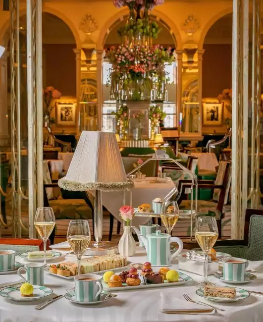 Claridge's otelinde lüks bir öğleden sonra çayı deneyimi sunan masada, pastel mavi çizgili porselen takımlar, zarif şampanya kadehleri, çeşitli atıştırmalıklar ve tatlılarla dolu tepsiler, arka planda görkemli iç mekan detayları yer almaktadır.