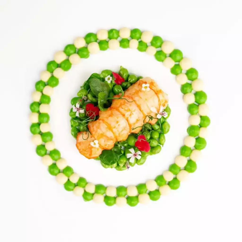 Core by Clare Smyth restoranından, merkezinde parlak turuncu karideslerin taze yeşil bezelye, yapraklar ve zarif yenilebilir çiçeklerle çevrelendiği, beyaz tabak üzerinde açık yeşil ve krem rengi küçük toplardan oluşan dairesel bir halka ile süslenmiş, sanatsal ve göz alıcı bir deniz mahsülü sunumu.