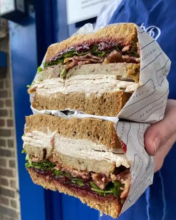 De Beauvoir Deli'nin menüsünden olduğu anlaşılan, bol malzemeli ve ortadan ikiye kesilmiş bu iştah açıcı sandviçte, kalın kahverengi ekmek dilimleri arasında bolca dilimlenmiş hindi, çıtır pastırma, taze yeşillikler ve koyu kırmızı kızılcık sosu katmanları göz alıcı bir şekilde sergileniyor.