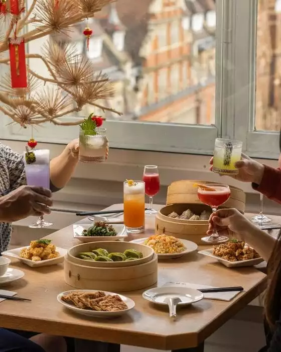 Din Tai Fung restoranında, Çin fenerleri ve kırmızı süslemelerle dekore edilmiş pencere kenarındaki bir masada, kişiler ellerinde çeşitli renklerde kokteyllerle kadeh kaldırırken, bambu buharda pişirme sepetlerinde yeşil mantılar, dim sumlar, kızarmış pilav ve farklı lezzetler sergileniyor.