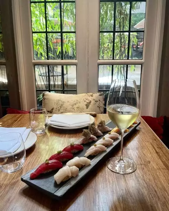 Dinings SW3 restoranının bambu ağaçlarının göründüğü pencere kenarında, ahşap bir masanın üzerinde şık bir sunumla taze ton balığı, deniz tarağı ve farklı balık türlerinden oluşan çeşitli nigiri sushi parçaları ile beyaz şarap dolu bir kadeh sergileniyor.