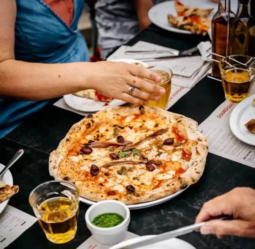 Bir Franco Manca restoranında çekilen bu karede, masanın merkezinde iştah açıcı, bol peynirli, zeytinli, kaparili ve ançuezli ekşi mayalı pizza dururken, etrafında arkadaşlarıyla keyifli bir yemek deneyimi yaşayan insanların elleri ve içecekleri görülüyor.