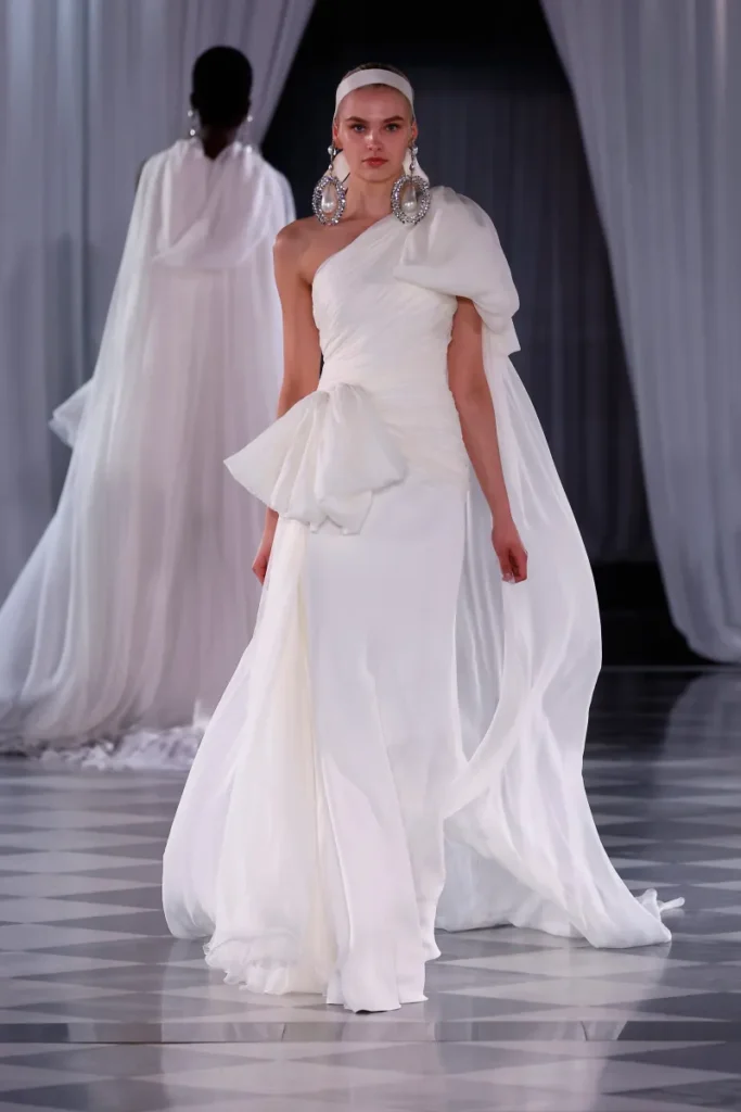 Giambattista Valli tasarımı, tek omuzlu ve dramatik drape detaylı gelinlik modeli. Eteğinde ve omuz düşüşünde zarif akışkanlık sunan bu tasarım, büyük fiyonk ve dev küpe aksesuarlarıyla teatral bir zarafet yansıtıyor.