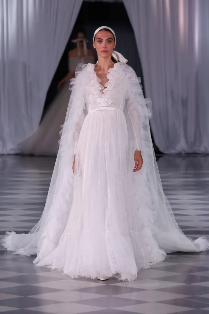 Giambattista Valli tasarımı, fırfırlı yakası ve uzun tül pelerin detaylarıyla öne çıkan romantik beyaz gelinlik. Hafif dokuya sahip, vintage esintili bu model, zarif ve nostaljik bir bridal görünüm sunuyor.