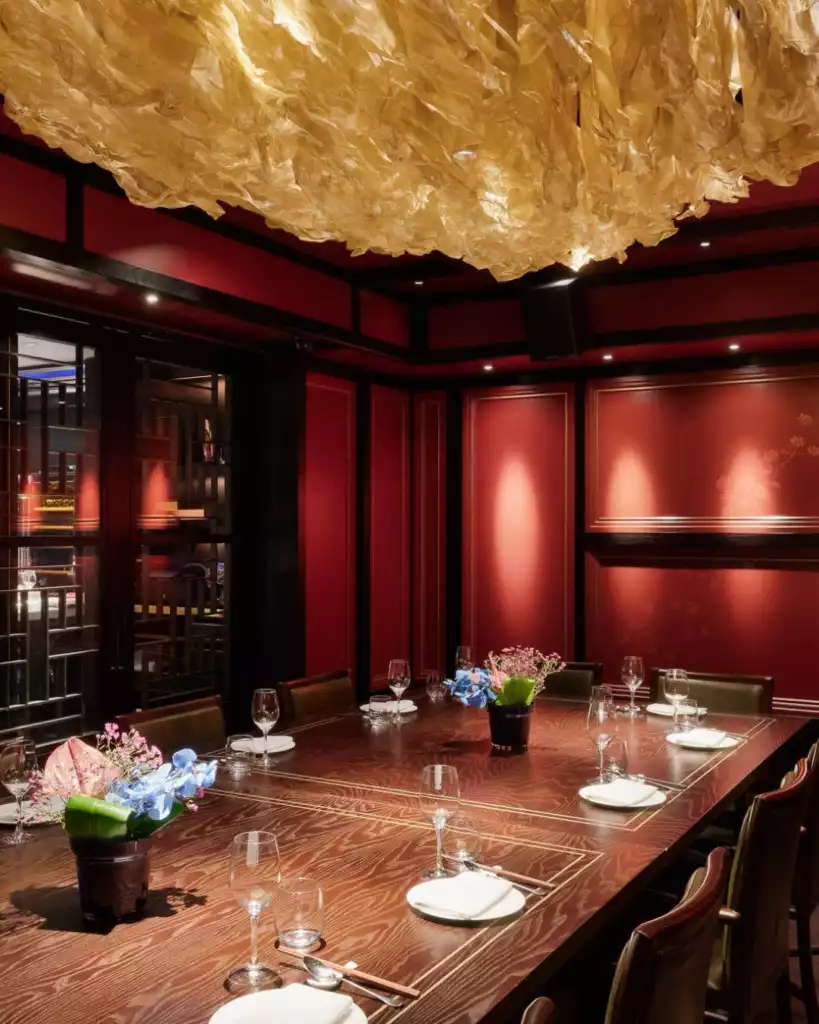 Hakkasan restoranındaki kırmızı duvarlı, koyu ahşap panelli, altın renkli avizeli özel yemek odası, masada zarif çiçek düzenlemeleri ve eksiksiz servis takımlarıyla lüks ve davetkar bir atmosfer sunuyor.