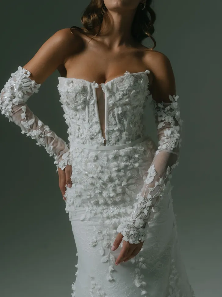 Hera Couture tasarımı, 3D çiçek aplikeleriyle süslenmiş, transparan uzun kollu ve kalp yaka detaylı zarif gelinlik modeli. Romantik ve sofistike çizgiler taşıyan bu tasarım, floral doku sever gelinler için ideal.