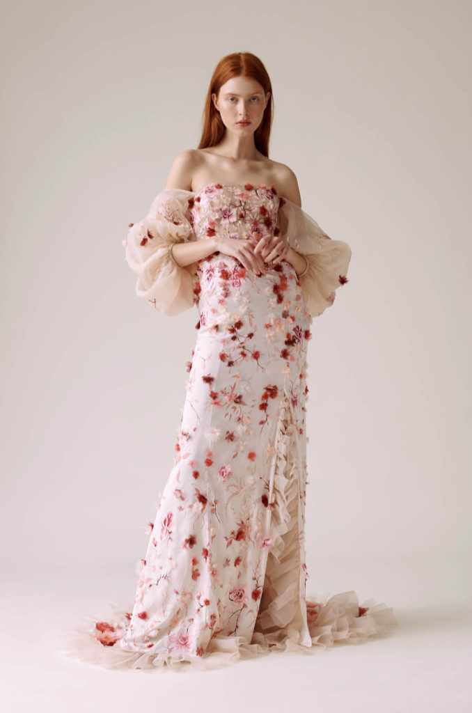 Hermione de Paula Atelier tasarımı, pastel tonlarda çiçek nakışlarıyla bezenmiş, düşük omuzlu ve kabarık transparan kollarla tamamlanan romantik gelinlik modeli. Zarif fırfır detayı ve bohem havasıyla farklılaşan sanatsal bir bridal görünüm sunuyor