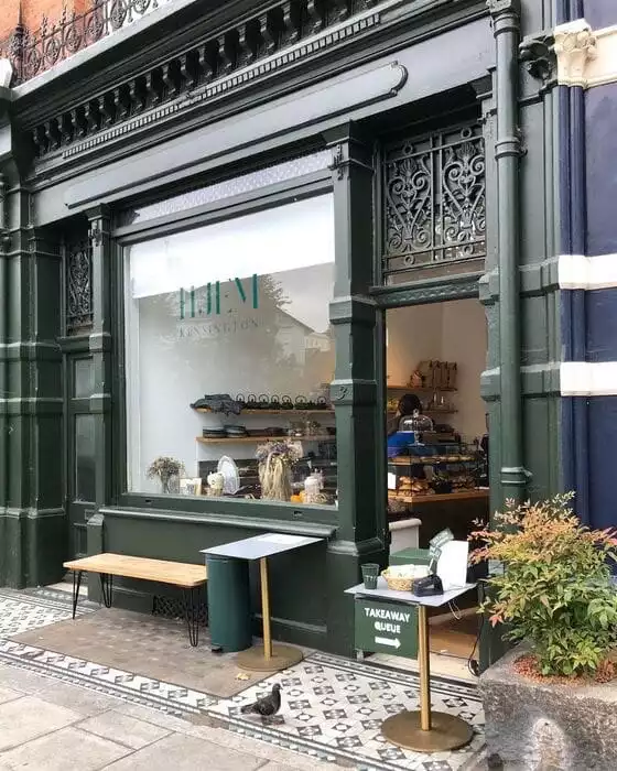 Koyu yeşil cepheli Hjem Kensington dükkanının önünde, fayans döşeli kaldırımda, ahşap bir bank, iki küçük masa, bir çöp kutusu ve üzerinde TAKEAWAY QUEUE yazan bir tabela bulunurken, vitrinde çeşitli ürünler sergileniyor ve bir güvercin kaldırımda yürüyor.