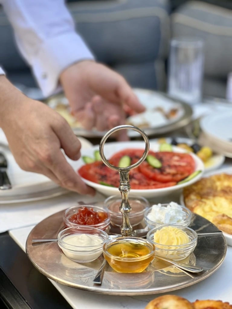 Gümüş renkli bir servis tepsisinde vişne reçeli, çikolata kreması, süzme bal, kaymak ve iki çeşit beyaz peynir/krema küçük cam kaselerde sunulmuşken, arka planda beyaz gömlekli bir kişinin masaya dilimlenmiş domates ve salatalık içeren bir tabak koyduğu görülüyor.