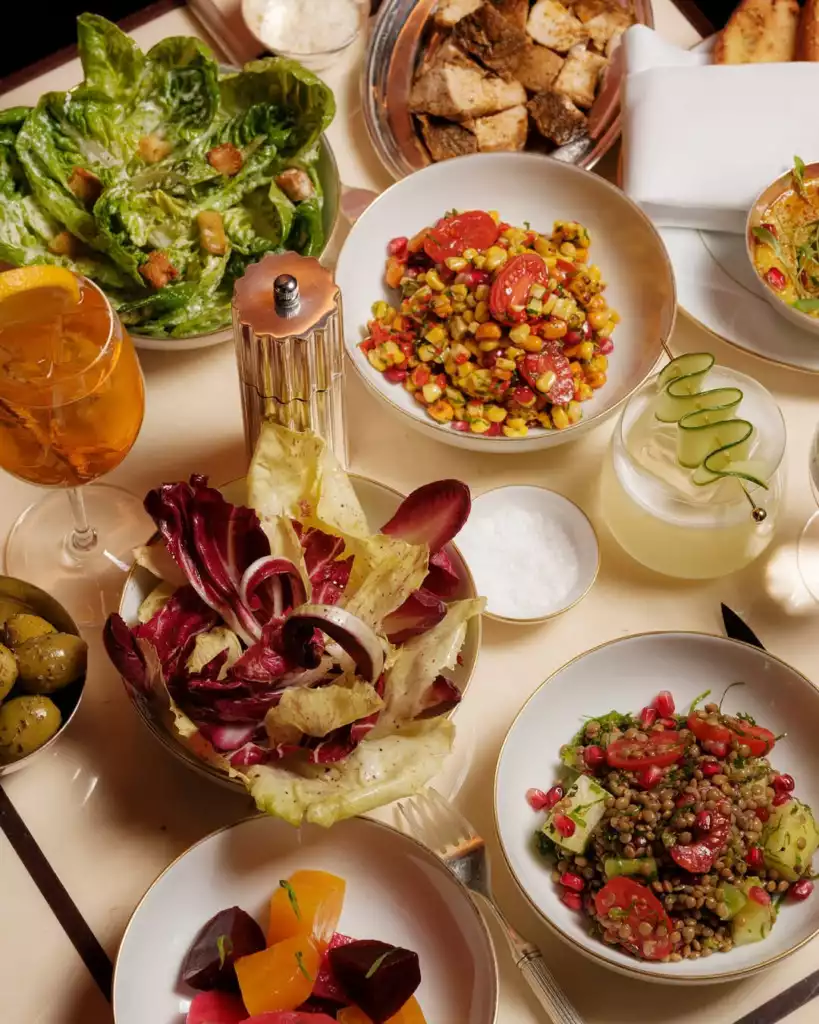 Bir masanın üzerinde taze yeşilliklerle hazırlanan Sezar salatası, mısır, nar ve domatesli mercimek salatası, kırmızı ve sarı pancarlı sebzeler, zeytinler ve ferahlatıcı içeceklerle dolu çeşitli renkli ve çekici meze ve salata kaseleri sergileniyor.