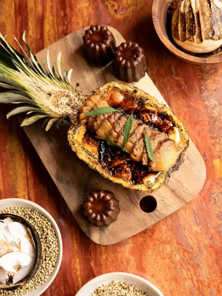 Ahşap bir kesme tahtası üzerinde, karamelize edilmiş ananas dilimleriyle dolu ve üzerinde taze yeşil yapraklar bulunan, dışı hafifçe kızarmış yarım ananas tatlısı; etrafında dört adet flüt şeklinde koyu kahverengi canelé tarzı minik kekler, sağ üstte tahta bir kasede ekmek ve sol altta hindistan cevizi parçacıkları ile karabuğday taneleri içeren kaselerle zengin bir sunum görülüyor.