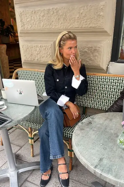 Matilda Djerf, dışarıda bir kafede otururken, denim ceket ve pantolonunu şık siyah babetlerle tamamladığı rahat ama özenli kombinasyonuyla kurumsal hayatta casual giyimin modern yorumunu etkili bir şekilde sergiliyor.