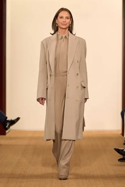 Bir manken, Ralph Lauren Sonbahar 2024 koleksiyonundan, kurumsal hayatta etkili giyinme için bej tonlarda uzun kruvaze bir ceket, uyumlu gömlek ve geniş paça pantolondan oluşan, profesyonel ve modern bir takım elbiseyi podyumda sergiliyor.