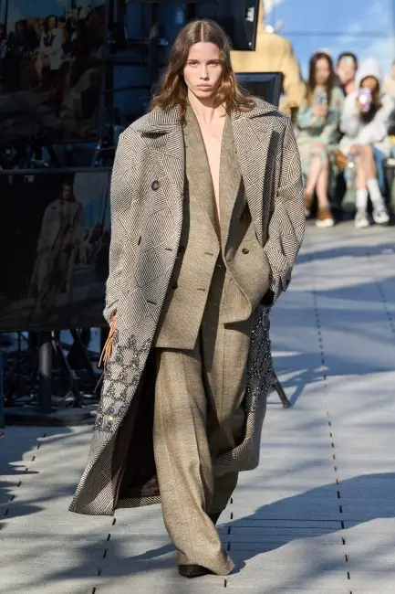 Uzun saçlı bir model, Stella McCartney defilesinde, kenarları ışıltılı yıldız desenleriyle süslenmiş gri desenli, oversized uzun bir palto ve altına giydiği bol kesim kahverengi takım elbiseyle podyumda yürüyerek kurumsal hayatta etkili giyim için modern ve sofistike bir yaklaşım sergiliyor.