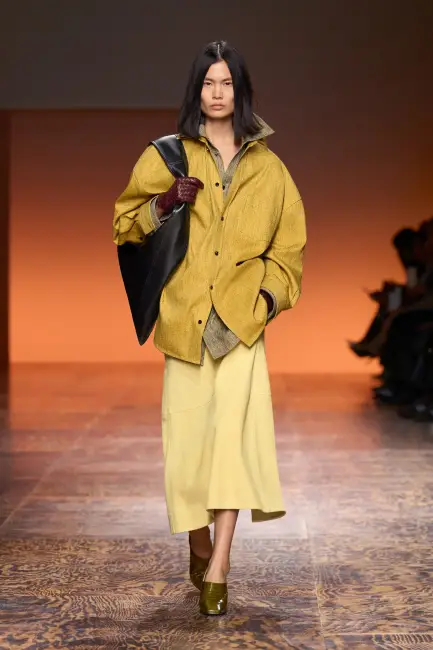 Bir model, oversized hardal rengi bir gömlek, açık sarı midi etek, bordo eldivenler, siyah omuz çantası ve zeytin yeşili topuklu ayakkabılarla podyumda yürüyerek, kurumsal hayatta hem şıklığı hem de rahatlığı dengeleyen etkili giyimin çağdaş bir örneğini sunuyor.