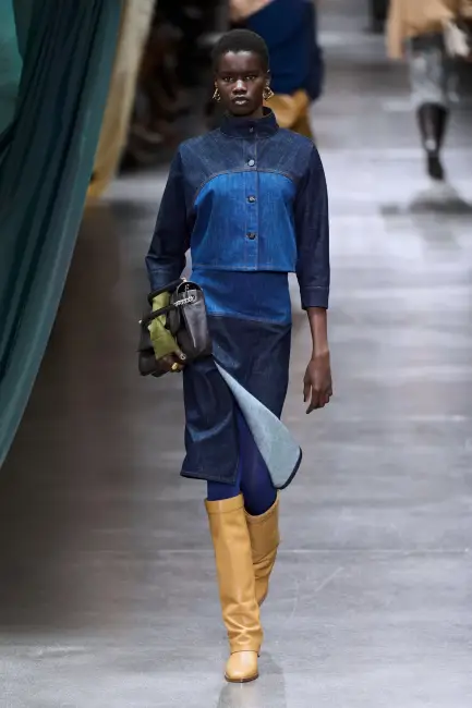 Podyumda yürüyen model, modern bir dokunuşla tasarlanmış iki tonlu denim ceket ve yırtmaçlı midi etekten oluşan casual bir kombinle, kurumsal hayatta özgün ve rahat bir stilin mümkün olduğunu gösterirken, görünümünü camel rengi yüksek çizmeler ve siyah bir el çantasıyla tamamlıyor.