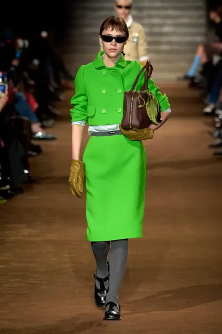 Model, Miu Miu'nun 2024 Sonbahar/Kış koleksiyonundan, canlı ve modern bir kurumsal stil sergiliyor; limon yeşili kısa kruvaze ceketi ve uyumlu kalem eteği, kahverengi şık çantası, koyu renk eldivenleri ve Mary Jane ayakkabılarıyla dikkat çekerek kurumsal hayatta etkili giyimin rahat ve şık yorumunu yansıtıyor.
