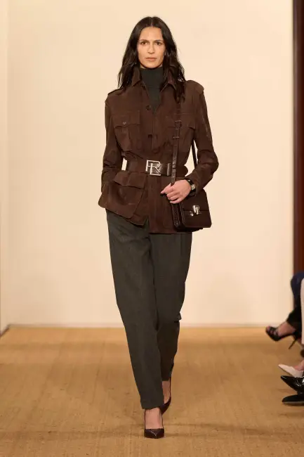 Ralph Lauren 2024 Sonbahar/Kış koleksiyonundan koyu kahverengi, kemerli, cep detaylı ceketi, zarif koyu gri bol kesim pantolonu ve uyumlu sivri burunlu topuklu ayakkabılarıyla podyumda yürüyen model, kurumsal hayatta modern ve etkili giyimin güçlü bir örneğini sunuyor.