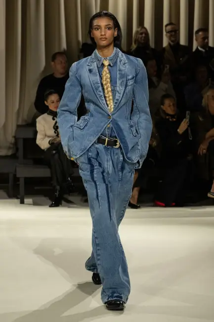 Kurumsal Hayatta Etkili Giyinme bağlamında, bir podyumda yürüyen model, belden oturan denim blazer ceket, pileli bol paça denim pantolon ve sarı dokulu kravat ile tamamlanmış denim gömlekten oluşan, Schiaparelli 2024 Sonbahar/Kış koleksiyonuna ait özgün bir jean takım elbiseyi sergiliyor; bu görünüm, casual giyim kodunda otoriter ve modern bir imaj sunarken, denim ve kurumsallık arasındaki dengeyi başarıyla yansıtıyor.