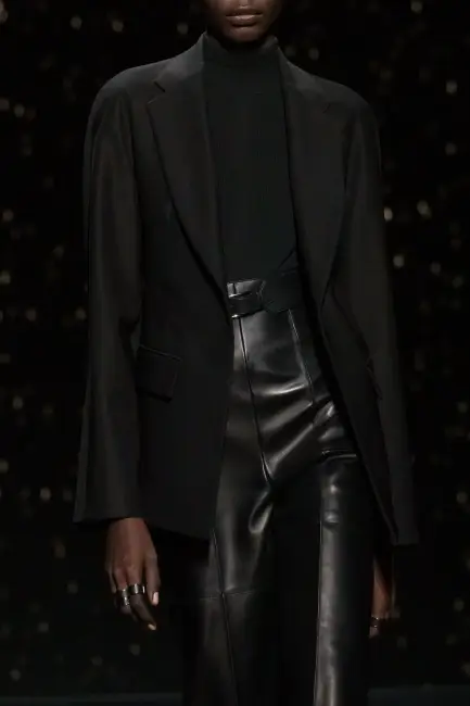 Koyu tenli bir model, siyah balıkçı yaka trikosu, rahat kesim blazer ceketi ve parlak siyah deri pantolonuyla, karanlık fonda modern bir duruş sergileyerek kurumsal hayatta etkili giyimin zarif ve çağdaş bir yorumunu sunuyor.