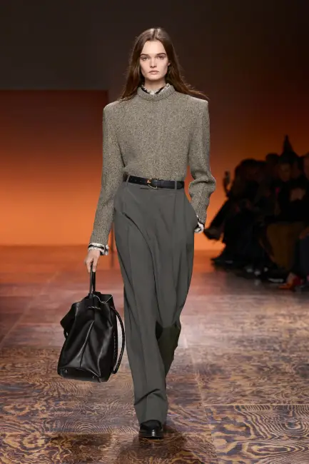 Bottega Veneta Sonbahar/Kış 2024 defilesinde, kabarık omuzlu bej-gri melanj bir kazak, siyah kemer, füme pileli bol paça kumaş pantolon ve siyah deri bir çanta taşıyan bir model, kurumsal hayatta etkili giyim için şıklığı konforla dengeleyen bir business casual kombiniyle podyumda yürüyor.