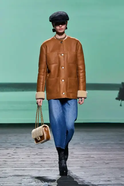 Mint yeşili zeminde yürüyen bir model, kahverengi içi kürklü deri ceketi, geniş kesim denim eteği, siyah topuklu botları, desensiz kasketi ve iki renkli küçük omuz çantasıyla kurumsal hayatta rahatlığın ve şıklığın birleşimini etkili bir şekilde sergiliyor.