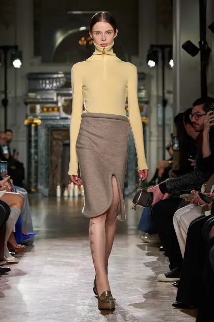 Victoria Beckham 2024 Sonbahar/Kış koleksiyonundan bir model, podyumda sarı yarım fermuarlı balıkçı yaka bluz, asimetrik kesimli, diz hizasında kum beji örgü etek, desenli şeffaf çoraplar ve altın rengi platformlu loafer ayakkabılar giyerek kurumsal hayatta modern, özgün ve etkili stilin cesur bir yorumunu sunuyor.