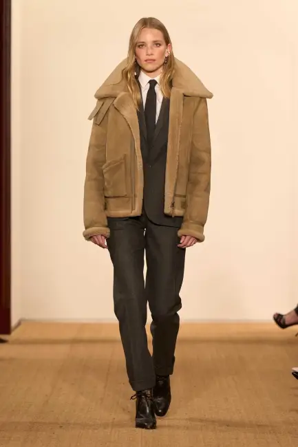 Ralph Lauren Sonbahar/Kış 2024 koleksiyonundan sarı saçlı bir model, koyu renk takım elbisesi, beyaz gömleği ve kravatıyla resmiyeti, üzerine giydiği kürklü kahverengi süet bomber ceket ve siyah bağcıklı botlarıyla harmanlayarak kurumsal hayatta etkili bir stilin modern ve özgün bir yorumunu sunuyor.