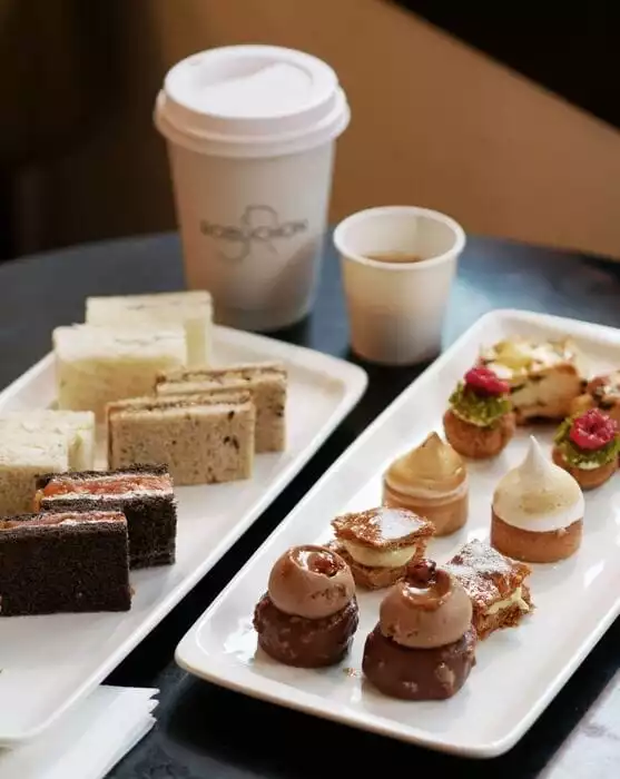 Le Deli Robuchon atmosferini yansıtan bu karede, solda ince kesilmiş sandviçler ve minik kekler, sağda ise kremalı, çikolatalı ve meyveli küçük pastacıkların yer aldığı iki beyaz servis tabağı ile arkada Robuchon logolu karton kahve bardağı bir arada zarifçe sunuluyor.