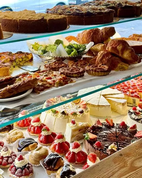Bir pastane vitrininde, çileklerle süslü bireysel tatlılar, çikolatalı eklerler, kat kat dizilmiş büyük pastalar, çeşitli kişler ve kruvasanlar gibi iştah açıcı yüzlerce farklı hamur işi özenle sergileniyor, bu da zengin bir menüyü işaret ediyor.