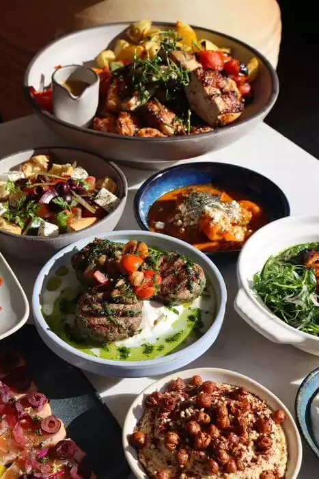 Bir masa üzerinde, ızgara tavuk parçaları, patates, zeytinyağlı salata, yoğurtlu köfte, humusun yanı sıra domates soslu karides gibi birçok farklı Akdeniz ve Orta Doğu yemeğinin sunulduğu çeşitli kaplar ve tabaklar görülüyor.
