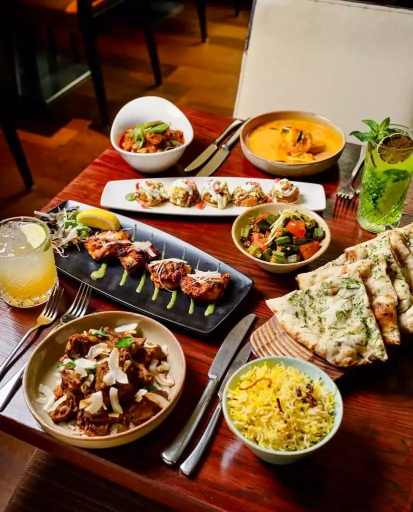 Mint Leaf restoranında sunulan, tandoori tavuk, zengin köri, baharatlı pilav, nan ekmeği, çeşitli mezeler ve nane yaprağı süslemeli ferahlatıcı kokteyllerle dolu, Hint mutfağının göz alıcı ve iştah açıcı bir ziyafet masası görüntüsü.