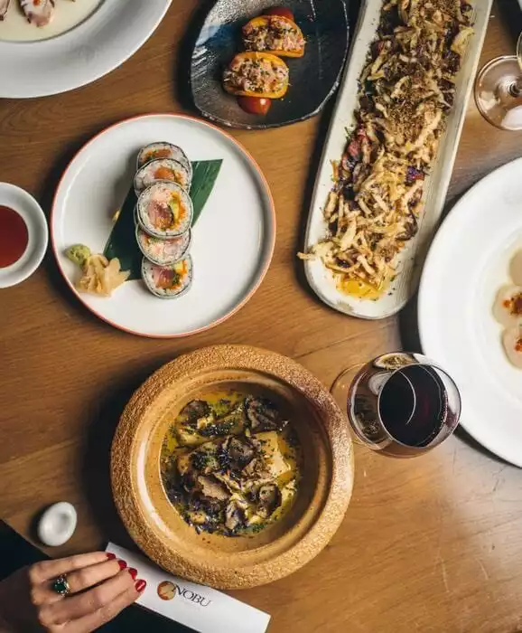 Nobu restoranında ahşap bir masa üzerinde, zarif suşi ruloları, çıtır soğanlı uzun bir meze tabağı, trüf mantarlı zengin bir ana yemek, diğer başlangıçlar ve kırmızı şarap kadehinin yer aldığı, Japon mutfağının özel lezzetlerini sergileyen davetkar bir ziyafetin kuşbakışı görüntüsü.