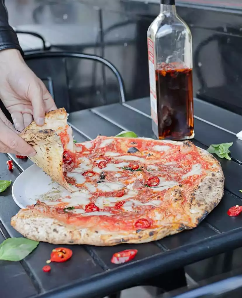 Siyah ahşap bir masada beyaz bir tabakta duran, üzeri taze domates sosu, erimiş mozzarella peyniri, yeşil fesleğen yaprakları ve parlak kırmızı biber dilimleri ile zenginleştirilmiş, kenarları odun ateşinde kızarmış lezzetli pizzadan bir dilimi kaldıran eller ve arka planda baharatlı yağ şişesi bir pizza deneyimini gösteriyor.