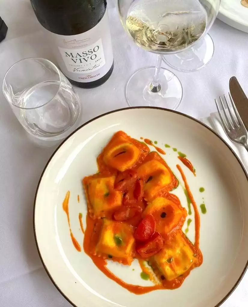 Beyaz masa örtüsü üzerinde, domates sosu ve kiraz domateslerle süslenmiş tabağın ortasında yer alan lezzetli ravioli, arka planda Masso Vivo Vermentino Frescobaldi beyaz şarap şişesi ve kadehi ile İtalyan mutfağının enfes bir deneyimini gözler önüne seriyor.