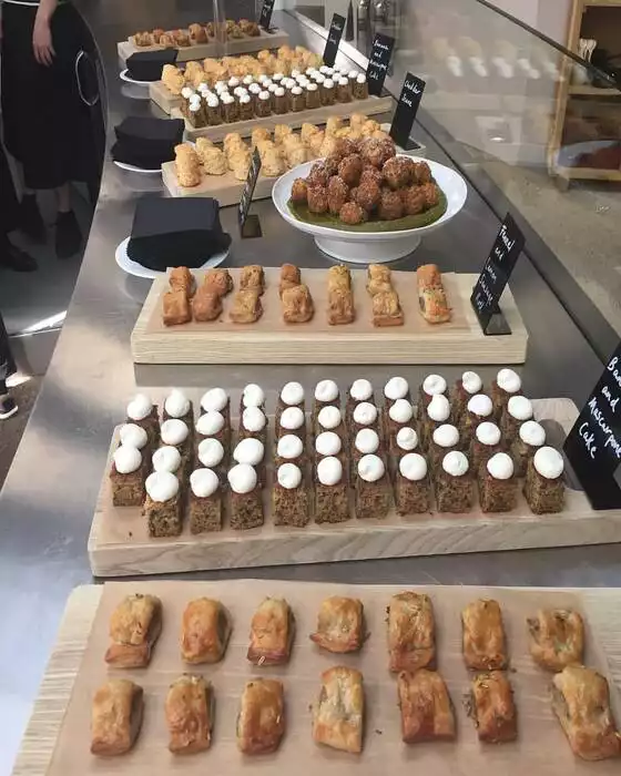 Paslanmaz çelik bir tezgahın üzerinde, bir Rose Fırını'nın sunduğu zengin açık büfe tarzı sergide, üzeri beyaz kremalı muzlu mascarpone kek dilimleri, alt sıradaki gevrek hamurlu mini sosis ruloları, çeşitli küçük tatlı ve tuzlu atıştırmalıklar ile yeşil bir sos yatağında özenle dizilmiş rezene ve limonlu sosis topları dikkat çekiyor.