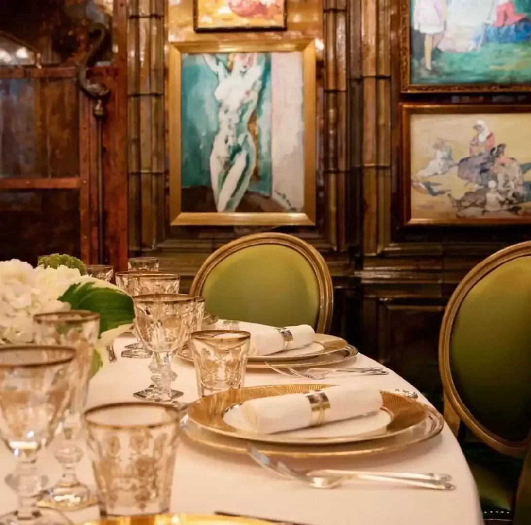 Scotts gibi lüks bir restoranda özel bir yemek deneyimi sunan, altın kenarlı zarif kadehler, altın rengi servis tabakları ve gümüş peçete halkalı keten peçetelerle özenle donatılmış, arka planında sanatsal tablolar ve yeşil kadife sandalyeler bulunan şık bir masa düzeni.