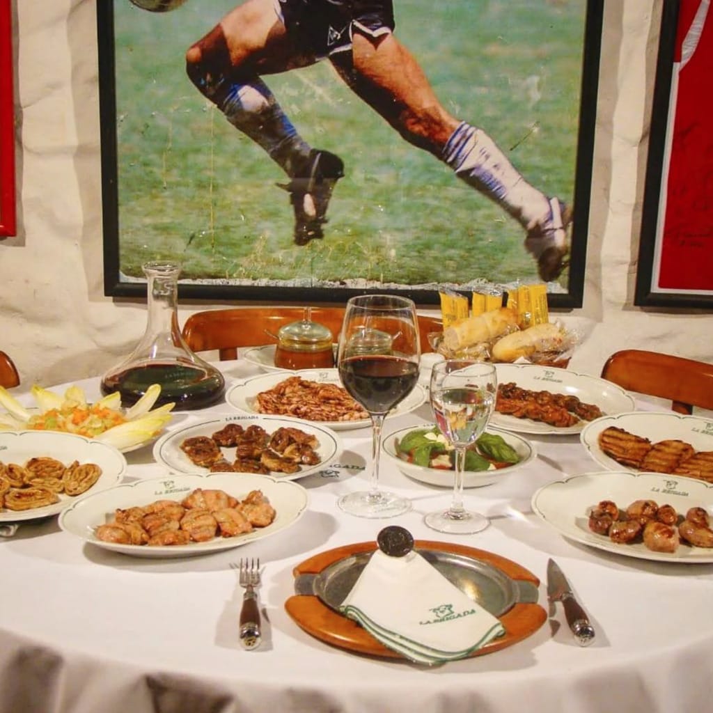 Arjantin et kültürünü yansıtan geleneksel bir parrilla olan La Brigada restoranında, bembeyaz bir örtüyle donatılmış yuvarlak masada servis edilen çeşitli ızgara etler ve şaraplar, arkada futbol temalı bir poster ile Buenos Aires'in lezzet dolu ve tutkulu atmosferini sergiliyor.