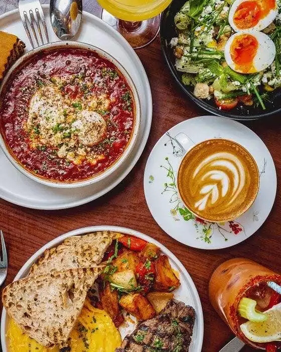 Bir Pazar brunch masasında, ortasında domates soslu fırınlanmış iki yumurta, haşlanmış yumurtalı salata, sütlü kahve, çırpılmış yumurta, biftek, fırın patates ve kızarmış ekmek gibi çeşitli lezzetler ile baharatlı bir içecek sunuluyor.