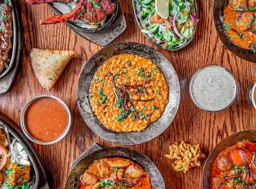 Bir ahşap masa üzerinde kuşbakışı sergilenen çeşitli Hint ve Pakistan lezzetleri; ortada kızarmış soğan ve taze yeşilliklerle zenginleştirilmiş büyük bir mercimek yemeği, yanında çıtır bir samosa, taze salata, parlak kırmızı soslu et yemekleri, yoğurtlu sos ve baharatlı güveçler ile dolu, iştah açıcı bir sofra görüntüsü.