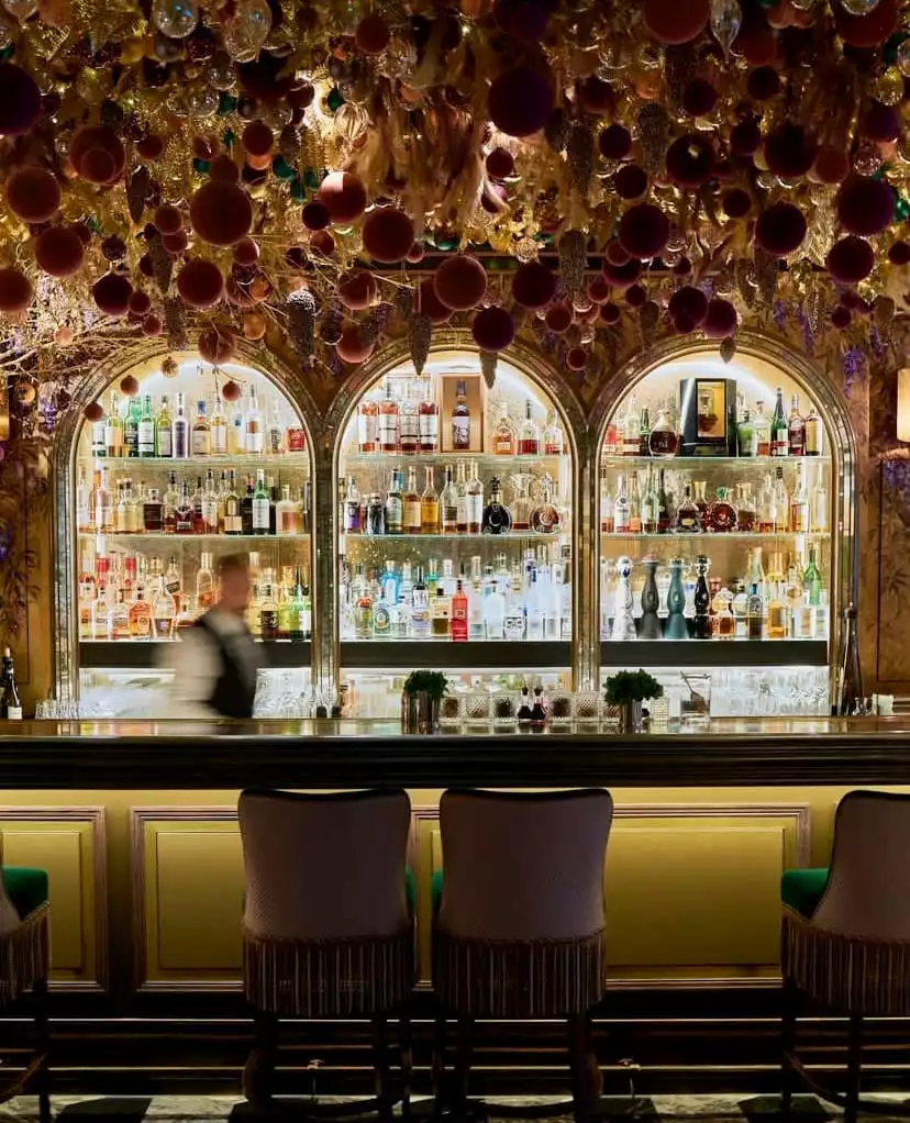 The Arts Club'ın zengin dekorasyonlu barında, altın rengi dallar ve bordo süs toplarıyla bezenmiş tavandan sarkan gösterişli düzenlemenin altında, arkasında bir barmenin hareket ettiği ışıklı içki şişeleriyle dolu üç kemerli raf ve önünde şık bar sandalyeleri yer alıyor.