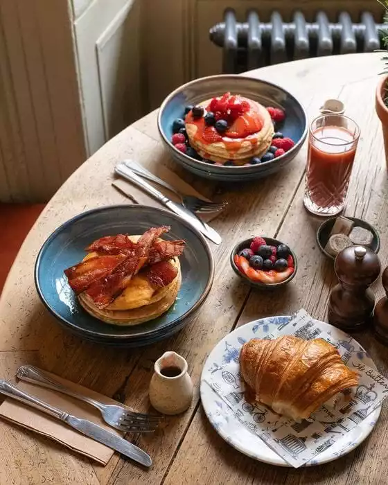 Ahşap bir masanın üzerinde, The Buttery'nin zengin kahvaltı menüsünden çilek ve yaban mersini ile süslenmiş pankekler, çıtır pastırmalı diğer pankekler, parlak altın rengi bir kruvasan, taze karışık meyve kasesi ve bir bardak içecekle dolu, iştah açıcı bir sofra düzeni görülüyor.