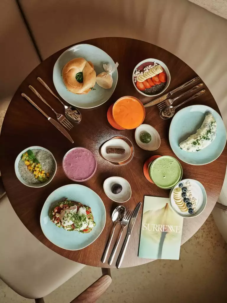 Yuvarlak ahşap bir masada, simit, çilek ve muz dilimli acai kasesi, avokado tost, chia pudingi, rengarenk smoothieler, meyveli yoğurt ve Surrenne adlı bir kitapla birlikte sunulan, zengin ve sağlıklı bir brunch kahvaltı düzenlemesi üstten çekim bir açıyla görülüyor.