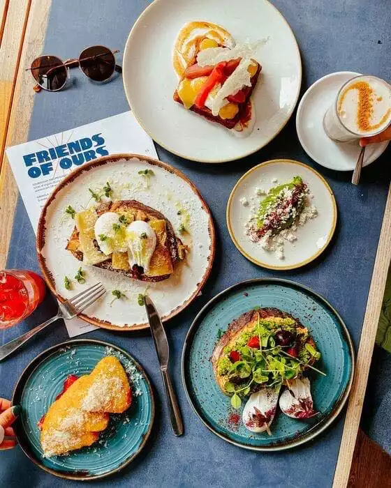 Friends of Ours kafesinde, mavi tonlarda bir masanın üzerinde özenle düzenlenmiş, çeşitli lezzetlerle dolu bir brunch ziyafeti sunuluyor: poşe yumurtalı ve rendelenmiş peynirli tost, kırmızı meyveler ve krema ile süslenmiş tatlı ekmek, pestolu ve taze yeşillikli somun ekmekler, pudra şekerli kızarmış hamur işleri ve avokadolu küçük bir meze tabağı. Yanında sıcak kahve, serinletici içecekler ve güneş gözlüğü ile keyifli bir öğün deneyimini yansıtan zengin bir görsel.