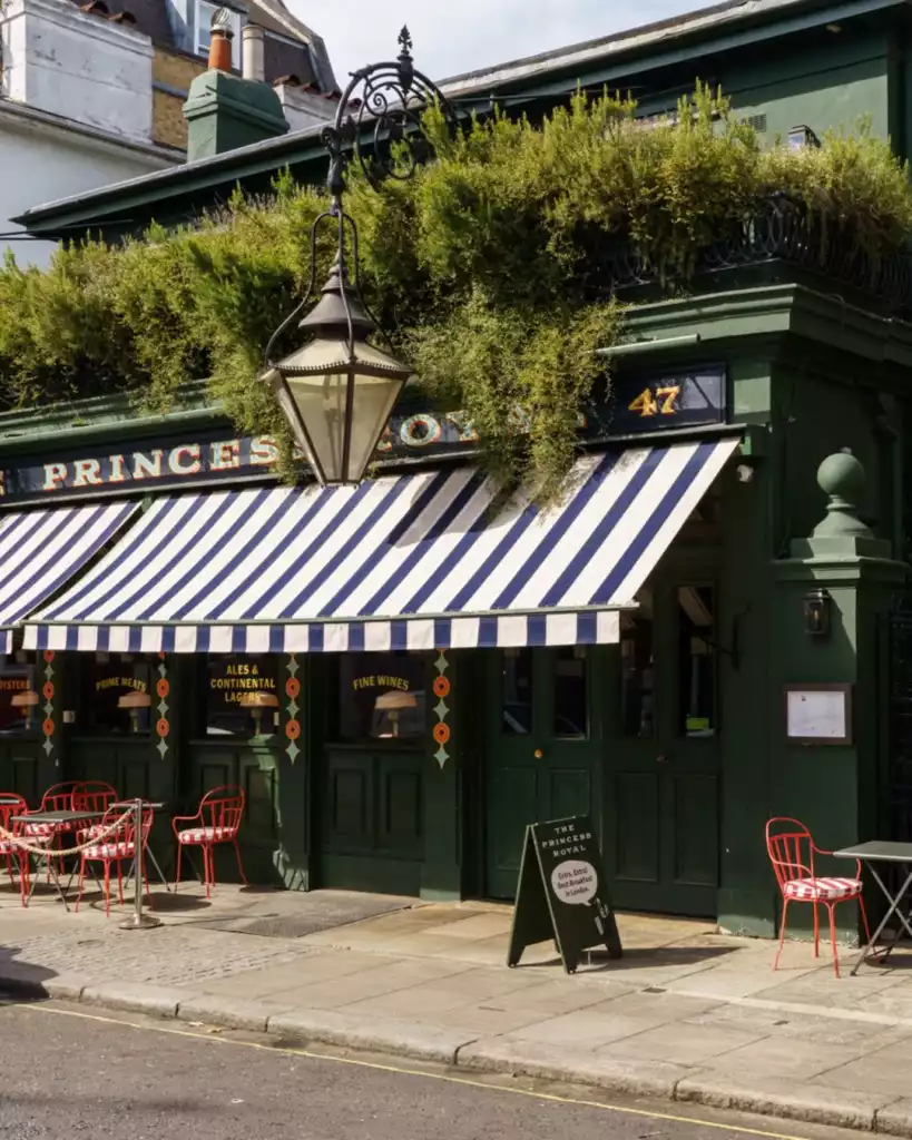 Londra'daki The Princess Royal adlı mekanın, koyu yeşil dış cephesi, mavi ve beyaz çizgili tenteleri, çatısını süsleyen yeşillikleri ve önünde kaldırıma dizilmiş kırmızı masa ve sandalyeleriyle sokak manzarasına hakim, eski tarz bir pub veya restoran atmosferi sunan çekici görüntüsü.