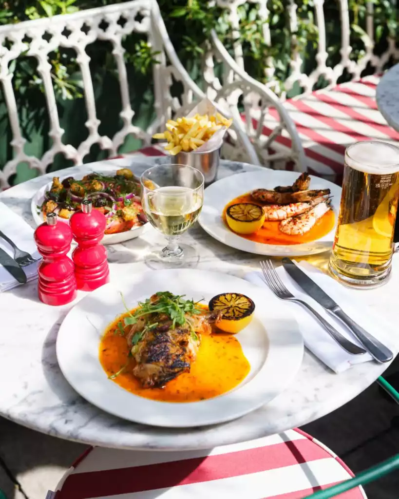 Güneşli bir dış mekan restoranında, mermer masa üzerinde servis edilmiş, portakal soslu ızgara tavuk ve karides tabakları, yanında patates kızartması, taze sebzelerle dolu bir salata, kadehte beyaz şarap ve bira bulunan davetkar bir yemek görünümüyle arka planda bitki örtüsü ve süslü beyaz sandalyeler.