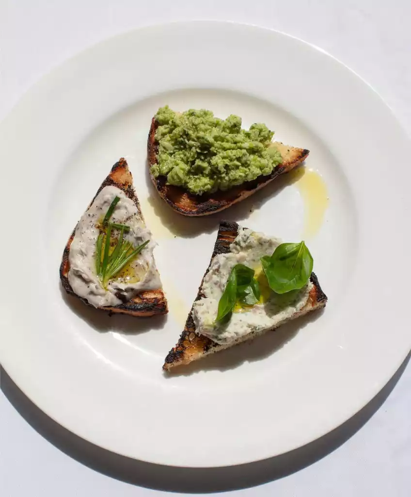 The River Cafe'nin sunduğu nefis başlangıçlardan biri olan bu karede, beyaz bir tabakta özenle sunulmuş üç farklı lezzetteki kızarmış ekmek dilimi (bruschetta) yer alıyor; biri parlak yeşil bezelye ezmesiyle, diğeri biberiye ve zeytinyağı gezdirilmiş kremalı beyaz bir karışımla, üçüncüsü ise taze fesleğen yaprakları ve zeytinyağı ile taçlandırılmış benzer bir ezmeyle servis ediliyor.