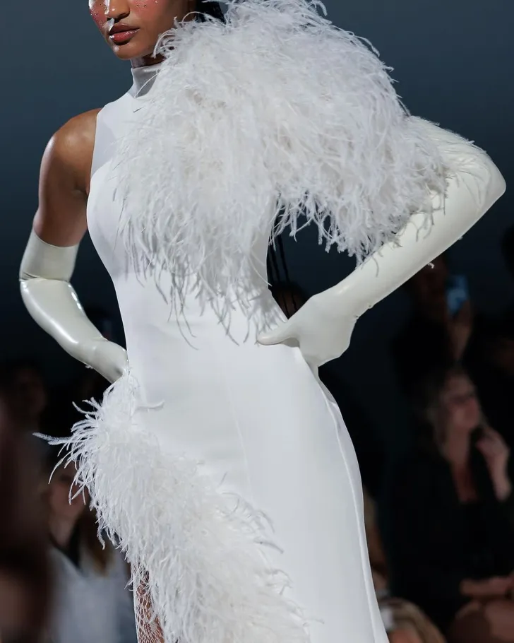 WONA Concept imzalı, beyaz saten kumaş üzerine tüy detaylarıyla süslenmiş modern gelinlik. Tek omuz ve etek kısmındaki dramatik kuş tüyü dokunuşlarla göz alıcı bir couture şıklığı sunuyor. Uzun beyaz eldivenlerle tamamlanan zarif ve iddialı bir siluet.