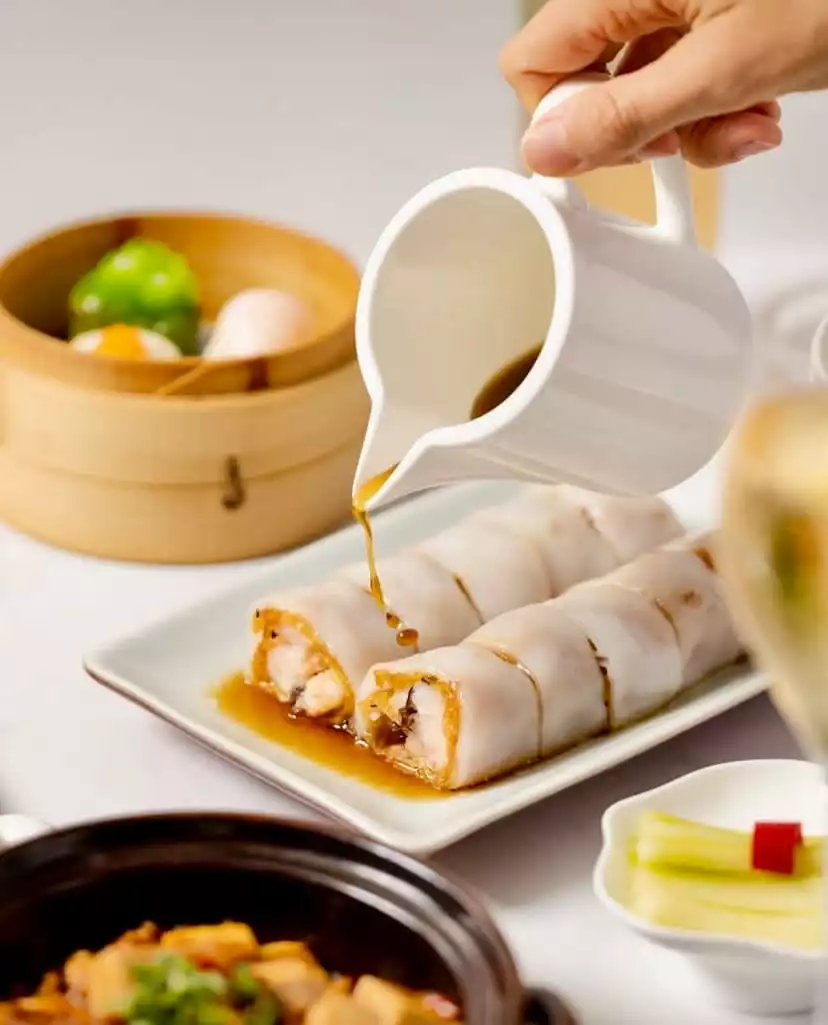 Bir el, beyaz bir tabağın üzerindeki iki parlak pirinç noodle rulosuna (cheung fun) özenle koyu kahverengi sos dökerken, arka planda bambu buharda pişiricideki çeşitli dim sumlar ve masadaki diğer Çin lezzetleri iştah açıcı bir sunum oluşturuyor.