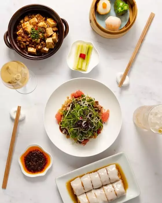 Yauatcha'nın menüsünden şık bir sunumla beyaz mermer bir masada üstten görüntülenen bu fotoğrafta, bambu bir buharda pişirici içinde göz alıcı dim sum çeşitleri, yanında bol yeşillikli ve greyfurtlu bir salata, güveçte baharatlı tofu yemeği, soya soslu pirinç erişte ruloları ve serinletici içecekler yer alıyor.